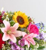 A vibrant and diverse floral arrangement featuring a large yellow sunflower, light pink lilies, a bright pink rose, and various purple and white accent flowers with green foliage.