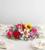 A lush and vibrant low-profile floral centerpiece titled "Painter’s Garden," featuring a mix of pink roses, a yellow sunflower, a red gerbera daisy, and pink lilies on a white dining table.