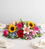 A vibrant and colorful horizontal floral centerpiece featuring sunflowers, pink lilies, red gerbera daisies, and hot pink roses on a white tablecloth.
