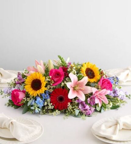 A vibrant and colorful horizontal floral centerpiece featuring sunflowers, pink lilies, red gerbera daisies, and hot pink roses on a white tablecloth.