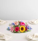 A vibrant, low-profile floral centerpiece titled "Painter's Garden," featuring a sunflower, pink lilies, hot pink roses, and blue delphiniums, arranged on a white tablecloth with four place settings.
