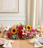 A vibrant floral centerpiece on a wooden dining table featuring sunflowers, pink lilies, red gerbera daisies, and hot pink roses, flanked by two lit taper candles.