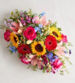 A vibrant and colorful floral centerpiece featuring pink lilies, yellow sunflowers, red gerbera daisies, and magenta roses mixed with small blue and white wildflowers and lush greenery.