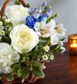A serene floral arrangement in a wicker basket featuring white roses, white carnations, blue delphiniums, and white alstroemeria, set against a warm wooden background with a blurred candle.