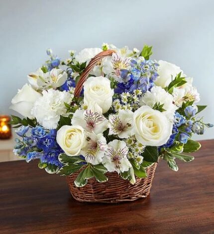 A lush floral arrangement in a brown wicker basket featuring white roses, white carnations, white alstroemeria, and vibrant blue delphinium, set on a wooden table.