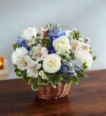 A serene floral arrangement featuring white roses, white carnations, blue delphinium, and alstroemeria in a classic brown wicker basket, placed on a dark wooden table.