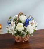 A serene floral arrangement in a small wicker basket featuring white roses, white carnations, blue delphinium, and white alstroemeria with purple accents, set on a dark wooden table against a light blue wall.