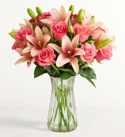 A lush bouquet of vibrant pink roses and light pink Asiatic lilies arranged in a clear fluted glass vase against a white background.