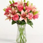 A lush bouquet of vibrant pink roses and light pink Asiatic lilies arranged in a clear fluted glass vase against a white background.
