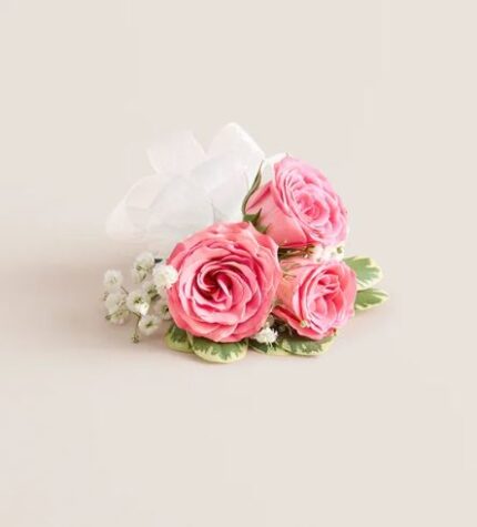 Three pink spray roses with white baby’s breath and a white satin ribbon bow, arranged as a wrist corsage on a light beige background
