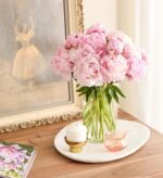 A lush bouquet of light pink peonies in a clear glass vase sits on a white marble tray atop a wooden table. Next to it are a white candle in a gold holder, a small pink glass vessel with matchsticks, and a framed painting of a ballerina in the background.