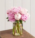 Lush bouquet of pink and white peonies with full, ruffled blooms in a clear glass jar vase, set on a rustic wooden table against a white paneled background.