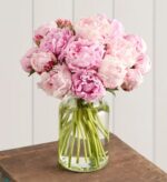 A lush bouquet of fully bloomed light pink and blush peonies in a clear glass vase, displayed on a rustic wooden table against a white paneled wall.