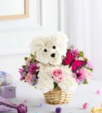 Floral arrangement shaped like a white puppy made from carnations, sitting in a wicker basket surrounded by pink roses, purple daisies, and pink chrysanthemums, with a pink butterfly accent and gift boxes in the background.