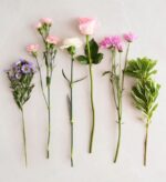 Six different flower stems arranged in a row on a light marble background, from left to right: purple and pink statice with small daisy-like blooms, pink carnations, white carnation, light pink rose, magenta chrysanthemums, and green variegated foliage.