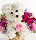 Floral arrangement shaped like a white puppy’s face made from white carnations, with black bead eyes and nose, surrounded by pink daisies, purple asters, a light pink rose, and a bright pink butterfly accent in a wicker basket.