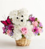 A floral arrangement shaped like a white puppy sits in a small wicker basket, crafted from white carnations and hydrangeas with black bead eyes and nose. The puppy holds a pink rose. It’s surrounded by pink roses, pink carnations, purple daisies, and fuchsia chrysanthemums. A bright pink and black butterfly decoration is perched on the left side.