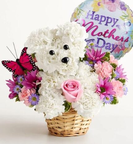 White floral puppy sculpture made from carnations with black bead eyes and nose, sitting in a woven basket surrounded by pink roses, purple daisies, and pink flowers. A pink butterfly decoration sits on the left and a "Happy Mother's Day" balloon floats behind.