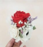 A preserved red rose boutonniere with a white rose, white anemone, baby's breath, and small lavender filler flowers, held between fingers against a soft beige background.