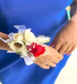 A red preserved rose boutonniere with white sola flowers, dried foliage, and light blue ribbon worn on a wrist against a royal blue dress.