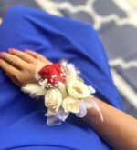 A close-up of a hand with coral nail polish resting on royal blue fabric, wearing a wrist corsage with one red preserved rose, three ivory preserved roses, white baby's breath, feathers, and sheer ivory ribbon.