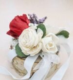 Preserved red rose and ivory sola wood roses with lavender sprigs, green leaves, and white satin ribbons arranged as a wrist corsage on a small birch wood slice base.
