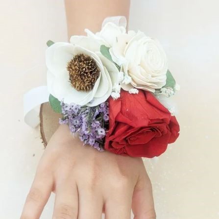 Wrist corsage on a hand featuring one red preserved rose, white sola wood flowers, small purple accent blooms, and greenery, tied with a white satin ribbon.