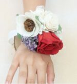 Wrist corsage on a hand featuring one red preserved rose, white sola wood flowers, small purple accent blooms, and greenery, tied with a white satin ribbon.