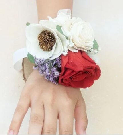 Wrist corsage on a hand featuring one red preserved rose, white sola wood flowers, small purple accent blooms, and greenery, tied with a white satin ribbon.