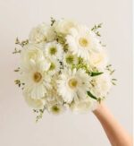 Hand holding a round bridal bouquet made of white gerbera daisies, white roses, and small white filler flowers with light green foliage, set against a plain off-white background.