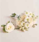 Elegant white prom bouquet with white roses, carnations, and chrysanthemums tied with white ribbon, paired with a matching single white rose boutonniere with green foliage, displayed on a soft beige background.