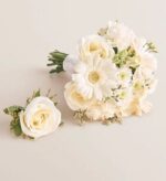 White prom bouquet and matching boutonniere on a light beige background. The bouquet features white gerbera daisies, roses, and hydrangeas with green accents, wrapped with white ribbon. The boutonniere is a single white rose with greenery.