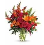 Vibrant deluxe flower bouquet in a clear glass vase with orange lilies, hot pink roses, orange spray roses, coral snapdragons, red carnations, purple accents, and green ferns.