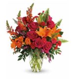 Vibrant floral bouquet in a clear glass vase featuring orange lilies, deep pink roses, red snapdragons, small red carnations, purple accent flowers, and green ferns.