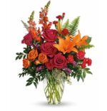 A vibrant floral arrangement in a clear glass vase featuring orange lilies, hot pink roses, orange spray roses, red carnations, snapdragons, purple accents, and green fern leaves.