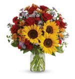 A vibrant mixed flower arrangement in a clear glass vase, featuring large yellow sunflowers, deep red roses, small orange daisies, and clusters of purple aster flowers with green foliage.