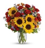 A vibrant mixed flower arrangement in a clear glass vase, featuring large yellow sunflowers, red roses, orange daisies, small purple aster flowers, and green foliage, set against a white background.