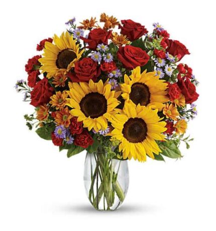 A vibrant mixed flower arrangement in a clear glass vase, featuring large yellow sunflowers, red roses, orange daisies, small purple aster flowers, and green foliage, set against a white background.