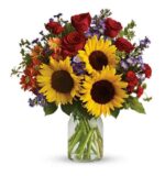 A vibrant mixed flower bouquet in a clear glass vase, featuring large yellow sunflowers, red roses, orange daisies, small red carnations, and purple accent flowers with green foliage.