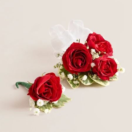 Red rose boutonniere with green leaves and floral tape next to a red rose wrist corsage with three roses, white baby's breath, variegated leaves, and sheer white ribbon, displayed on a light beige background