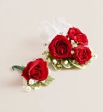 Red rose boutonniere with green leaves and floral tape next to a red rose wrist corsage with three roses, white baby's breath, variegated leaves, and sheer white ribbon, displayed on a light beige background