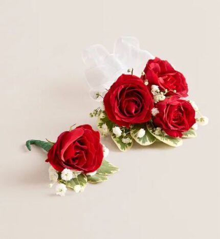 Red rose boutonniere with green leaves and floral tape next to a red rose wrist corsage with three roses, white baby's breath, variegated leaves, and sheer white ribbon, displayed on a light beige background