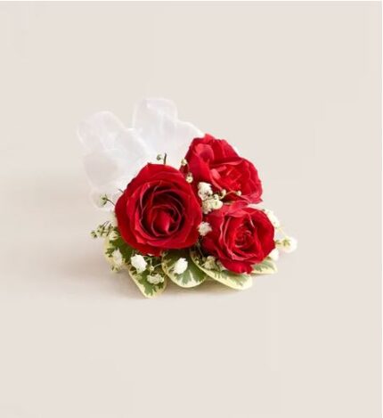 Red spray rose corsage with three deep red roses, white baby’s breath, variegated greenery, and white satin ribbon on a cream background.