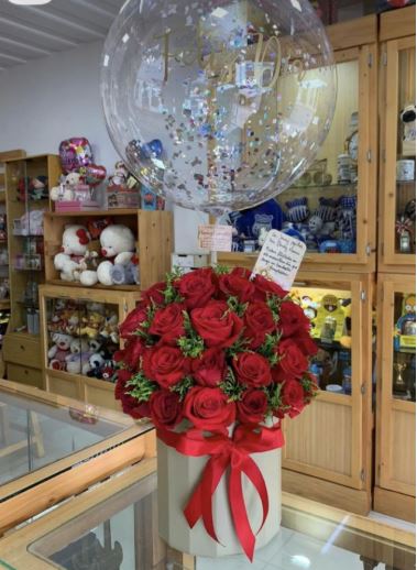 A luxurious floral gift arrangement featuring a large bouquet of red roses in a cylindrical grey box, topped with a clear bubble balloon filled with silver confetti.