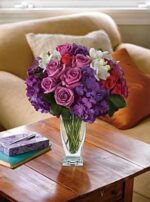 Elegant glass vase with purple hydrangeas, lavender roses, and white freesia on a wooden coffee table in a cozy living room