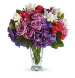 A lush floral arrangement in a clear glass vase featuring lavender roses, deep purple hydrangeas, red spray roses, white freesia, and green accents. The bouquet is full and rounded with rich purple, red, and white tones.
