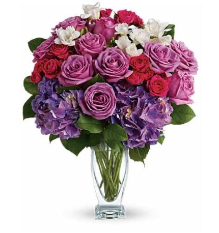 A lush bouquet of purple roses, red spray roses, purple hydrangea, and white freesia in a clear flared glass vase with visible green stems.