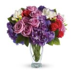 A lush bouquet in a clear glass vase featuring lavender roses, deep purple hydrangeas, red spray roses, white freesia, and light purple accents.
