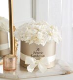 Round taupe hatbox with white ribbon and "Serene Gardenias" logo, filled with lush white preserved gardenias, styled on a marble tray next to a gold mirror and rose-gold candle.