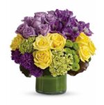 Round floral arrangement in a short green glass vase featuring yellow roses, purple roses, purple alstroemeria, green hydrangea, and purple carnations, accented with green button poms.